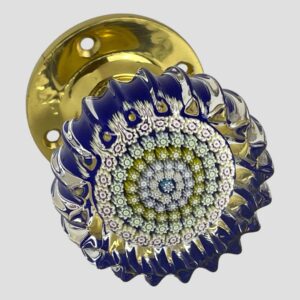 Main photo of Perthshire millefiori door knob shown with the crimped glass front in the right foreground with brass door fixing visible to the back top right corner. A blue millefiori is in the centre with letter P (for Perthshire) This is then following by rings of tightly packed different coloured millefiori pieces mostly in muted pastel shades. The base colour is cobalt blue with clear glass at the top end. The wavy crimpled edges all around give a feeling of the sea. The brass door fixing gives a quality finishing touch.