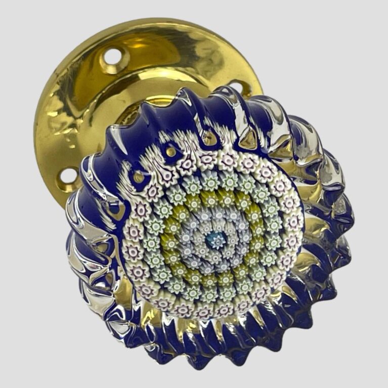 Main photo of Perthshire millefiori door knob shown with the crimped glass front in the right foreground with brass door fixing visible to the back top right corner. A blue millefiori is in the centre with letter P (for Perthshire) This is then following by rings of tightly packed different coloured millefiori pieces mostly in muted pastel shades. The base colour is cobalt blue with clear glass at the top end. The wavy crimpled edges all around give a feeling of the sea. The brass door fixing gives a quality finishing touch.