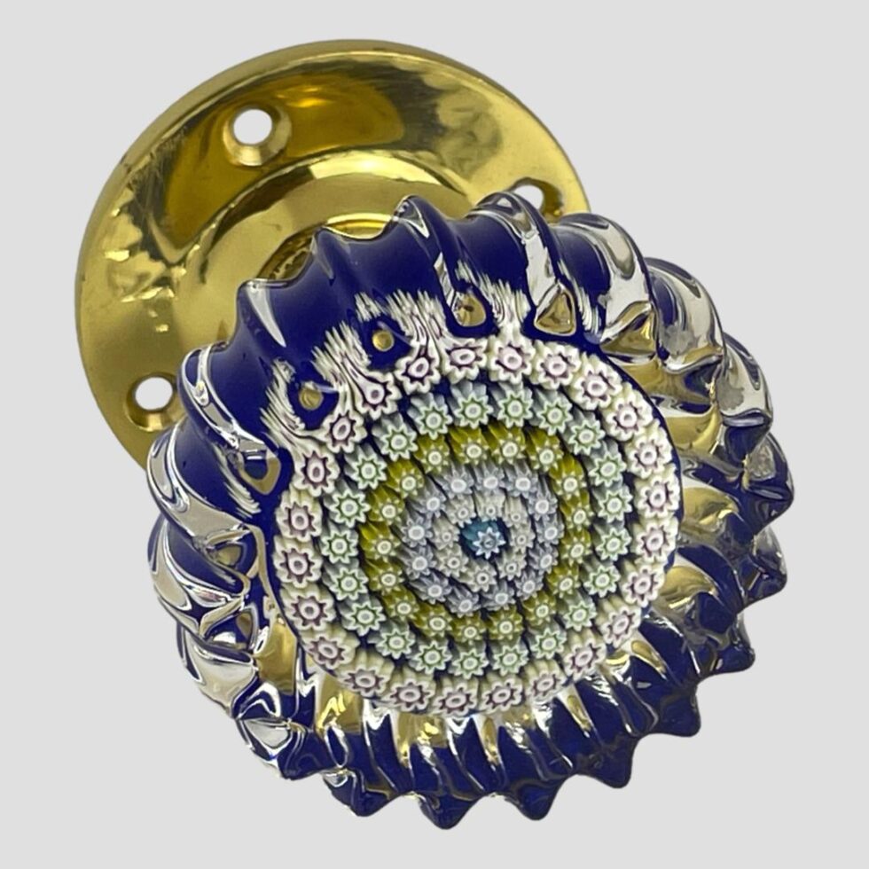Main photo of Perthshire millefiori door knob shown with the crimped glass front in the right foreground with brass door fixing visible to the back top right corner. A blue millefiori is in the centre with letter P (for Perthshire) This is then following by rings of tightly packed different coloured millefiori pieces mostly in muted pastel shades. The base colour is cobalt blue with clear glass at the top end. The wavy crimpled edges all around give a feeling of the sea. The brass door fixing gives a quality finishing touch.