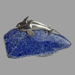 Main photo of Georg Jensen dolphins brooch displayed on top of a small rock of lapis lazuli. They are swimming side by side with one in front of the other. Bodies are arched as if in real swimming motion.