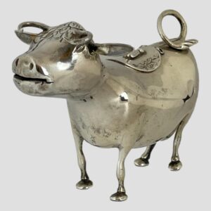 Main photo of Dutch silver cow creamer seen from an eye level with the cows head in the left foreground and curler over tail in the top right corner. The curled tail acts as the handle and the cows mouth is the spout all cleverly concealed! There is a hinged flap on the cows back.