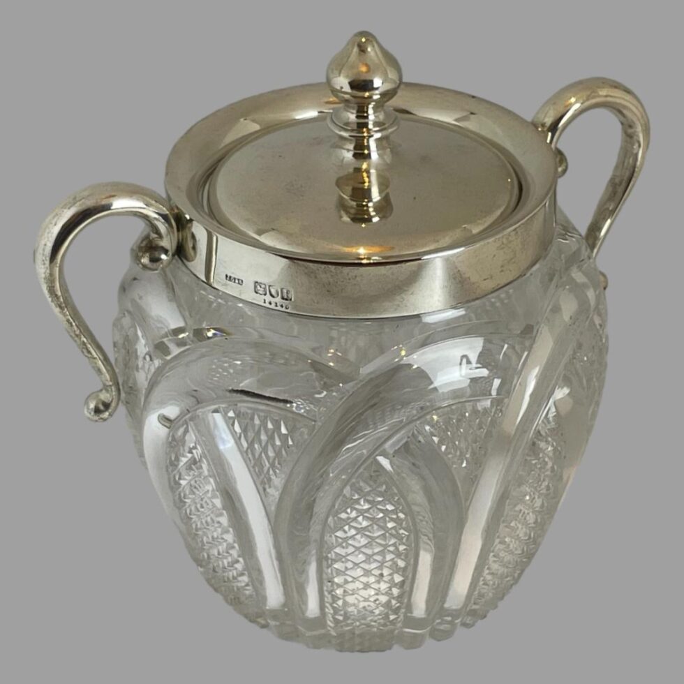 Photo of the glass &silver biscuit barrel seen this time from a little height looking down to the top of the lid diagonally. The barrel is set at an angle showing the hallmark to the left on rim.