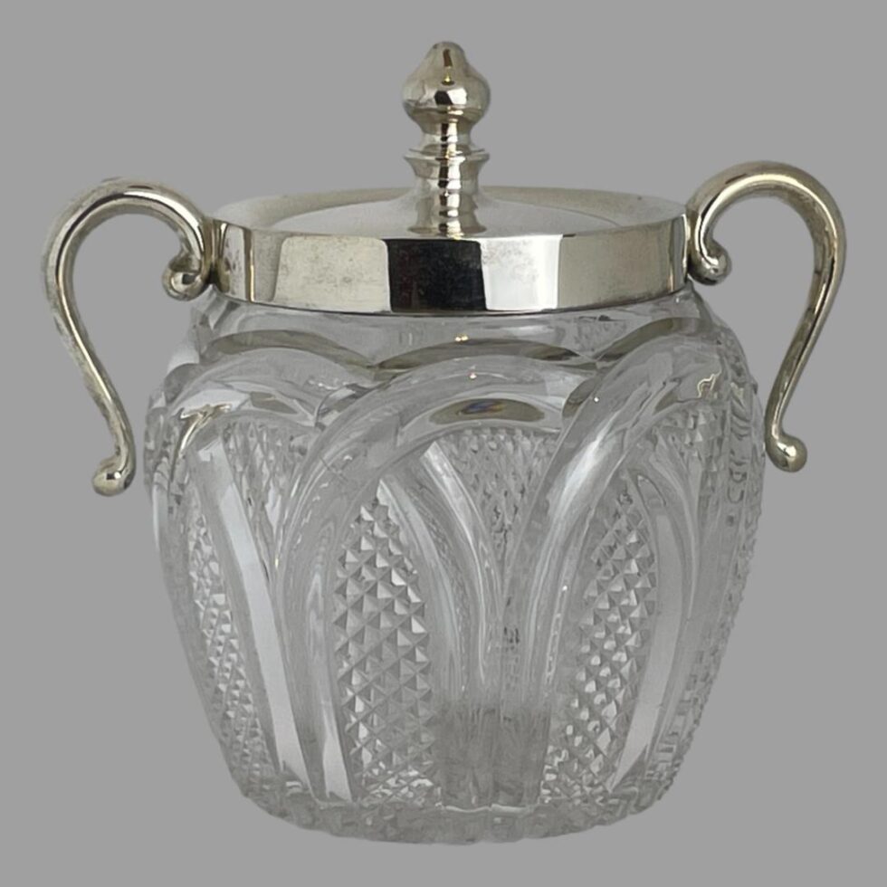 Main photo of glass &silver biscuit barrel seen from an eye level with the handles on left and right. The cut glass base is finely detailed which compliments that plain silver to the top. The silver handles have a upward loop with the ends hanging down to around the most bulbous part of the barrel. They are attached to the silver rim around the mouth. The lid also in silver has a fairly tall finial for easy removal. A lovely piece of late Victorian early Edwardian tableware.