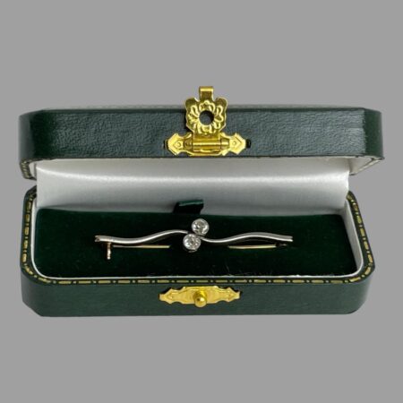 Main photo of Edwardian gold diamond brooch displayed inside its green case with the brooch front forward facing. The bar of the brooch has a twist effect in the middle where the 2 diamonds sit. The case has a gold coloured metal clasp closure and is included.