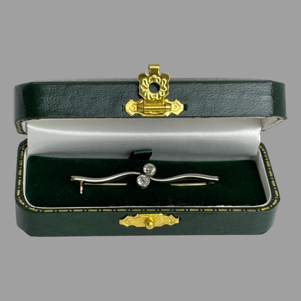 Main photo of Edwardian gold diamond brooch displayed inside its green case with the brooch front forward facing. The bar of the brooch has a twist effect in the middle where the 2 diamonds sit. The case has a gold coloured metal clasp closure and is included.