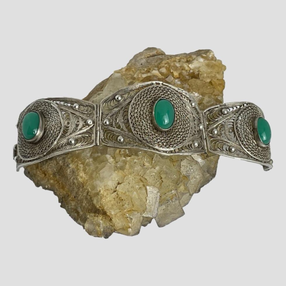 Main photo of Egyptian silver chrysoprase bracelet horizontally laid over a rock of river quartz from showing all 3 panels with vibrant green chrysoprase cabochon to the centre of each. The filigree silver work is exceptionally crafted. When the bracelet is closed, it becomes rigid like a bangle.