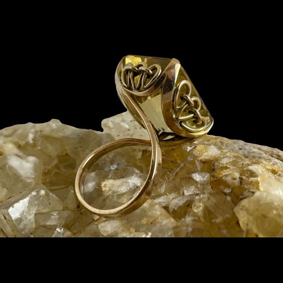 Photo of the large faceted citrine ring still displayed on top of a rock of river quartz this time with the ring front facing the top right corner to show the stone setting and the unusual band design. The framework holding the stone is also quite unusual with a sequence of loops and bars.
