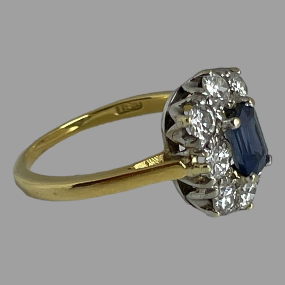 Photo of cornflower sapphire and diamond ring displayed on a flat surface with the ring front facing right. The gold hallmark is just visible to inside the band.