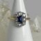 Main photo of cornflower sapphire diamond ring displayed on a white cone shaped stand with the ring front forward facing. The ring has a 'crown' design with one end slightly wider than the other - the wider worn at the top. The cornflower sapphire is a beautiful shade of blue with the 8 brilliant cut diamonds enhancing the overall look and quality of this ring. The stones are set in 18 karat white gold and the band is in yellow gold. There is a rock of natural quartz behind the cone display stand.