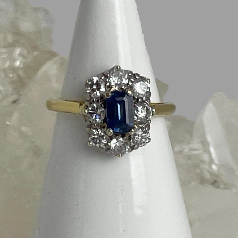 Main photo of cornflower sapphire diamond ring displayed on a white cone shaped stand with the ring front forward facing. The ring has a 'crown' design with one end slightly wider than the other - the wider worn at the top. The cornflower sapphire is a beautiful shade of blue with the 8 brilliant cut diamonds enhancing the overall look and quality of this ring. The stones are set in 18 karat white gold and the band is in yellow gold. There is a rock of natural quartz behind the cone display stand.