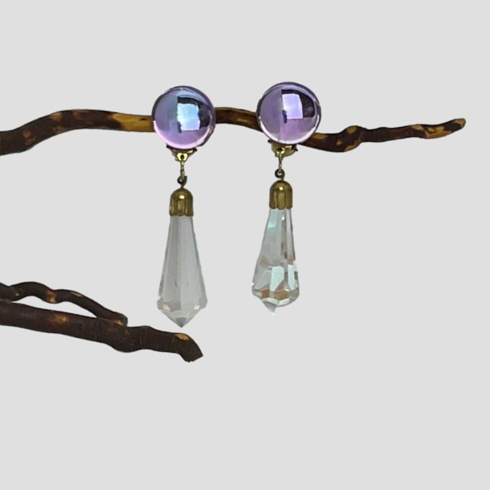 Main photo of vintage crystal clip-on earrings displayed hanging from a cherry twig and forward facing. The round crystals at top where the clips are attached are flashing purply blue while the hanging crystals appear clear with tiny hints of colour.