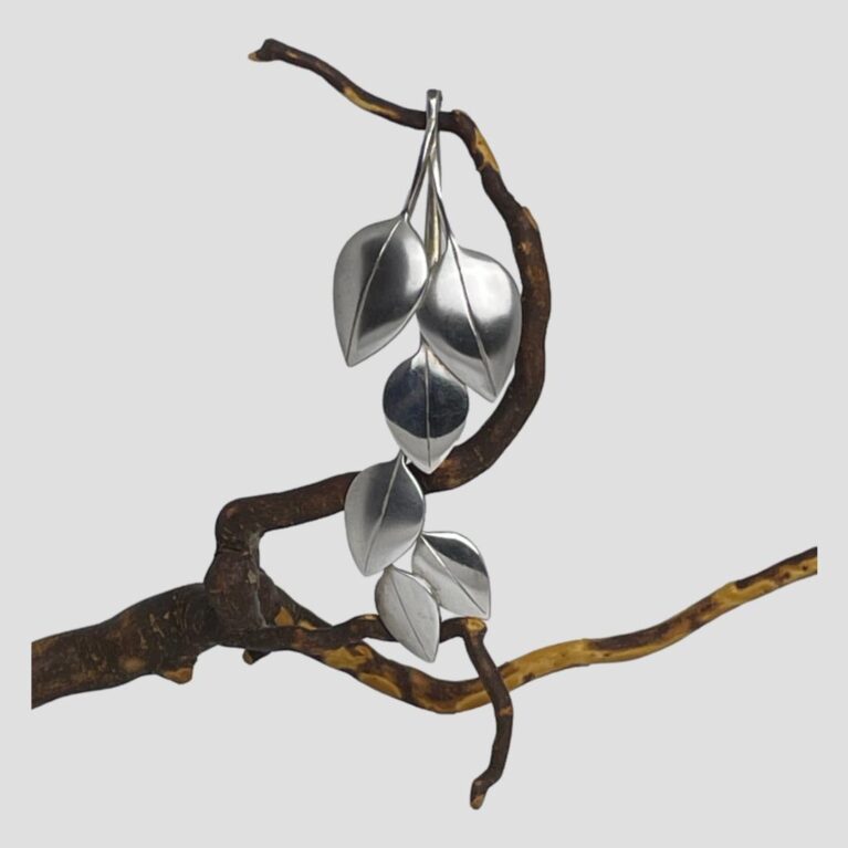 Main photo of silver articulated leaf brooch displayed hanging from a twig of dried cherry tree and looking straight on. The stem end at the top is hooked over a part of twig and the leaves hanging below. The articulated section of the brooch are the 3 leaves at the tip which are securely fastened with bars and loops. Very simple, stylish and elegant brooch.