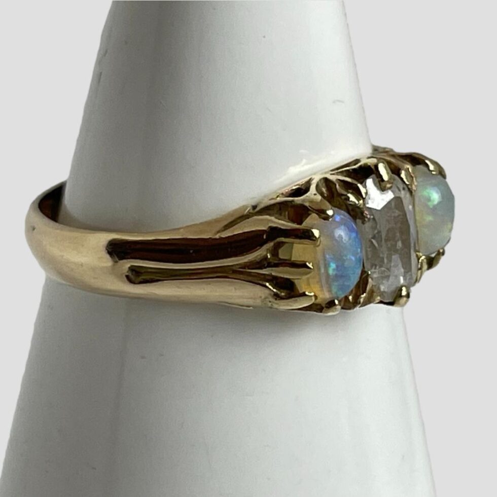 Photo of opal diamond ring displayed on a cone shaped stand with the ring front facing to the right. The debossed design on the shoulder appears like a trident.