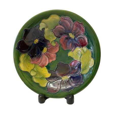 Main photo of large Moorcroft clematis bowl displayed on a dark wooden display stand (feet visible below bowl) The base colour of the bowl is in British racing green with a ring of clematis flowers and leaves covering most of the inside of bowl. The 4 flowers are in graduation from dusky pink to deep purple and the leaves are in a light green. This piece is signed by Walter Moorcroft, son to William.