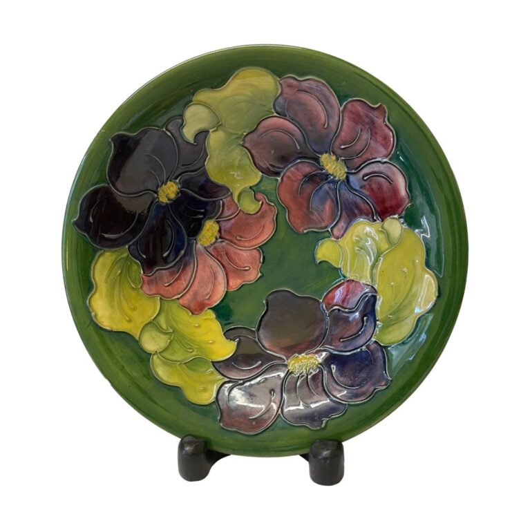 Main photo of large Moorcroft clematis bowl displayed on a dark wooden display stand (feet visible below bowl) The base colour of the bowl is in British racing green with a ring of clematis flowers and leaves covering most of the inside of bowl. The 4 flowers are in graduation from dusky pink to deep purple and the leaves are in a light green. This piece is signed by Walter Moorcroft, son to William.