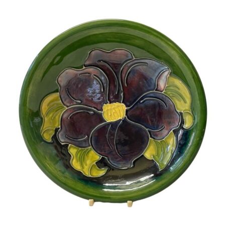 Main photo of Moorcroft clematis shallow bowl displayed upright on a plate stand with the inside of bowl on display. The bowl is in iconic British racing green with a large deep purple / magenta clematis flower at the centre with light green coloured leaves protruding out from behind the flower.