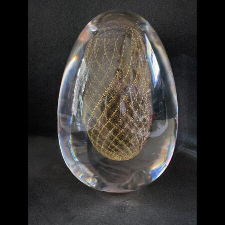 Main photo of vintage Murano egg paperweight displayed against a dark grey background. On the inside of the egg is a golden egg shape in net form so an egg shape made up of golden criss-crossed strands. The exterior of the egg is clear glass. A beautifully and expertly crafted piece.