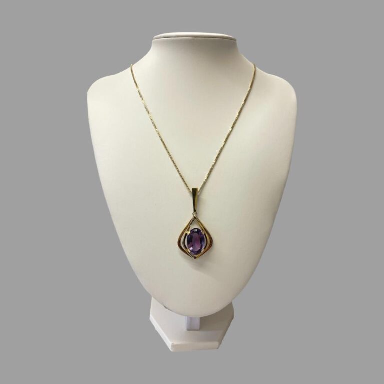Main photo of gold amethyst pendant &chain displayed on a necklace display stand of an off white colour and shown with the front of necklace facing forward. The pendant has a fairly long bail shaped like the top of an exclamation mark and the main shape of the pendant frame is a like a teardrop with a pointed tip at the end. There are 2 'frames' of gold around the large oval cut amethyst - the inner frame has 4 clips which hold the amethyst in place and which are positioned approximately at 2, 5, 7 and 10 o'clock. A second frame identical to inner frame just wider and longer, sits outside the inner frame. The chain is a nice box chain with good thickness which compliments the pendant perfectly.