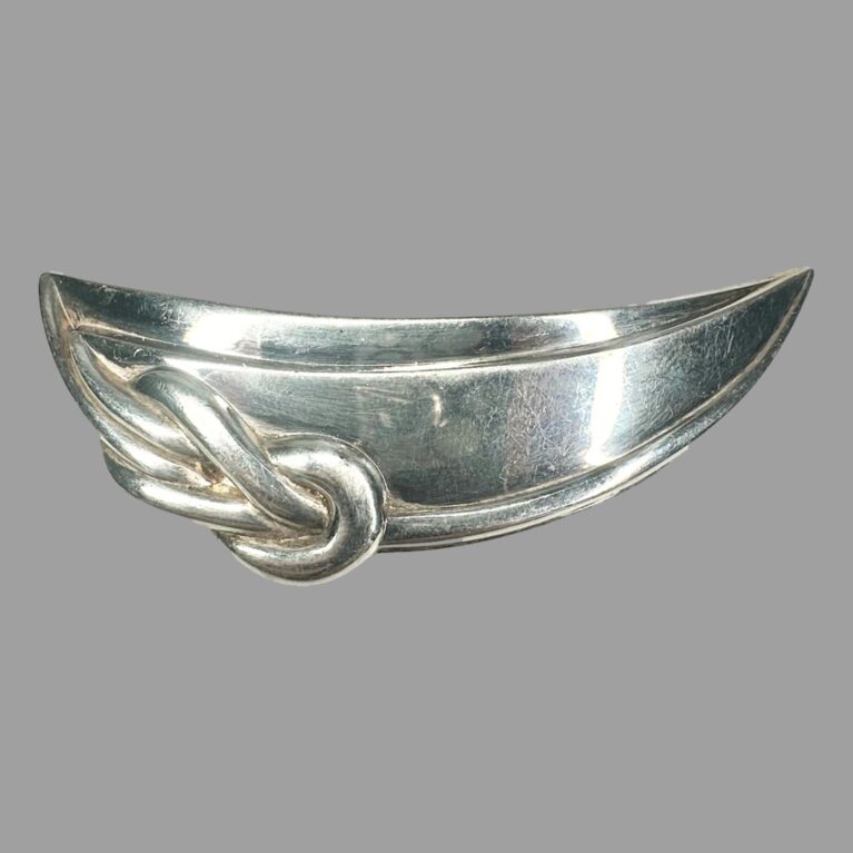 Main photo of Christofle silver plate brooch displayed with the brooch front forward facing. This brooch can of course be worn any way round. The brooch is shaped like a crescent with a reef knot detail to one rounded side on the left. On closure consideration, it could be a rowing boat shape hence reef know? The silver plate is in good condition.