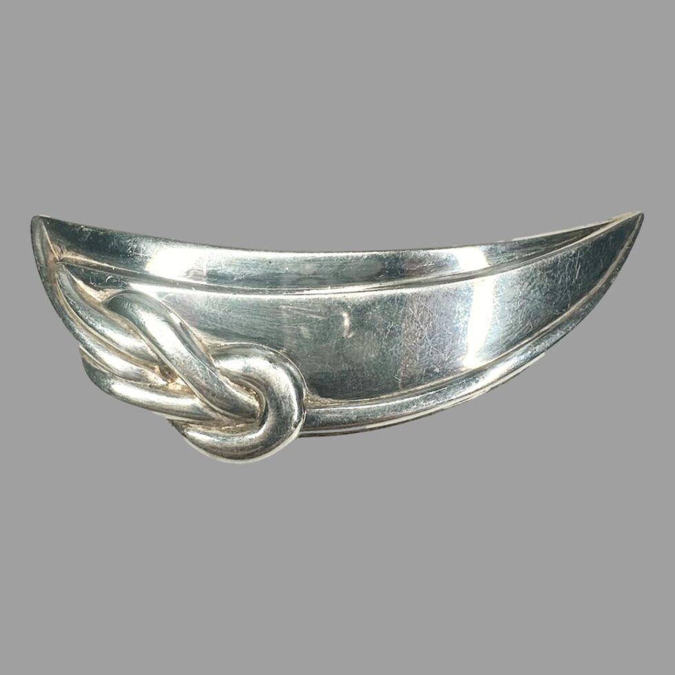 Main photo of Christofle silver plate brooch displayed with the brooch front forward facing. This brooch can of course be worn any way round. The brooch is shaped like a crescent with a reef knot detail to one rounded side on the left. On closure consideration, it could be a rowing boat shape hence reef know? The silver plate is in good condition.