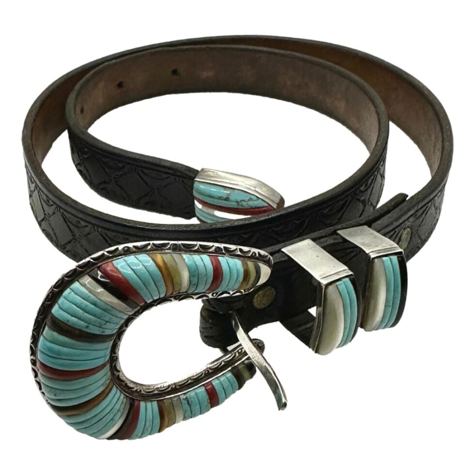 Main photo of Zuni inlaid silver belt displayed coiled like a snake for want of a better description! The buckle is in the foreground and shaped like a horseshoe with multiple stones inlaid mostly turquoise with other stone inlays set alternately. The belt tip is also inlaid as well as the 2 belt loops. The tip is at the centre of the coil. Finished with a tooled black leather strap. A statement belt if ever there was!