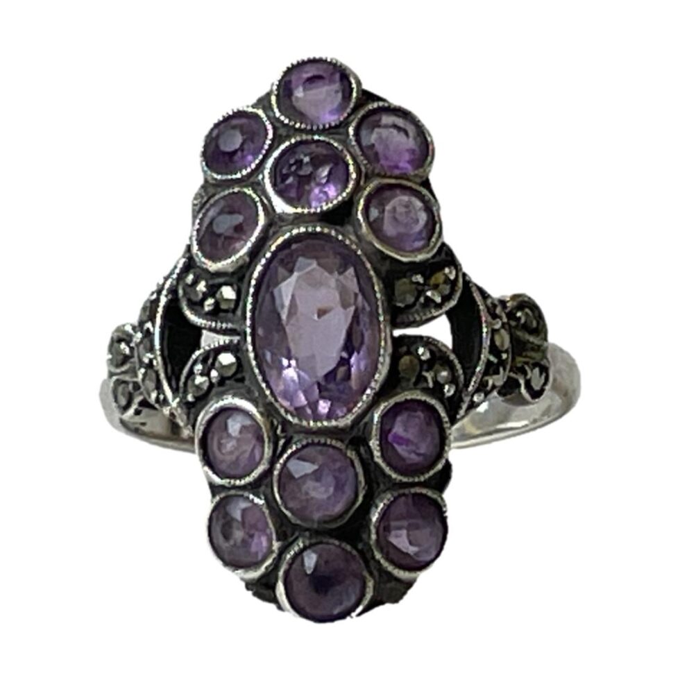 Photo of 935 silver &amethyst ring displayed on a flat surface with the ring front forward facing. The photo is in close up to show the individual setting of each stone in silver. Marcasite to left and right of central amethyst which is oval cut and set vertically. There is a small triangular gap then a few more marcasite on the shoulders. Lovely purple colour of the amethysts.