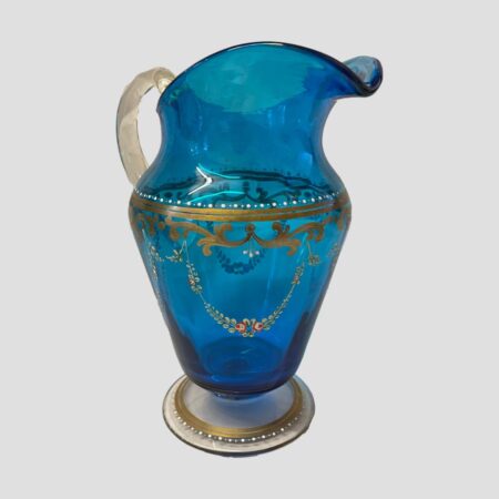 Main photo of antique Bohemian glass jug displayed with the spout facing to the bottom right corner and handle in top left. This is generous sized beautiful peacock blue coloured glass jug with a wide top and high spout. The handle is a clear glass as is the base rim. A lovely garland of small green foliage and small pink flowers are hand painted around the vase. They hang down from just below the gilt decoration around the neck area which is highlighted with white jewel dots above. The gilt continues around the base in 2 rings with further white jewel dots around the outer ring.