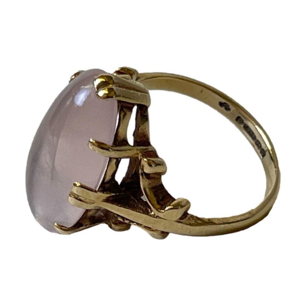 Photo of rose quartz ring displayed on a flat surface with the ring front facing left. The shot is taken from a slight angle looking down so the top and one side of setting is visible. The hallmark is also just visible on the inside band.