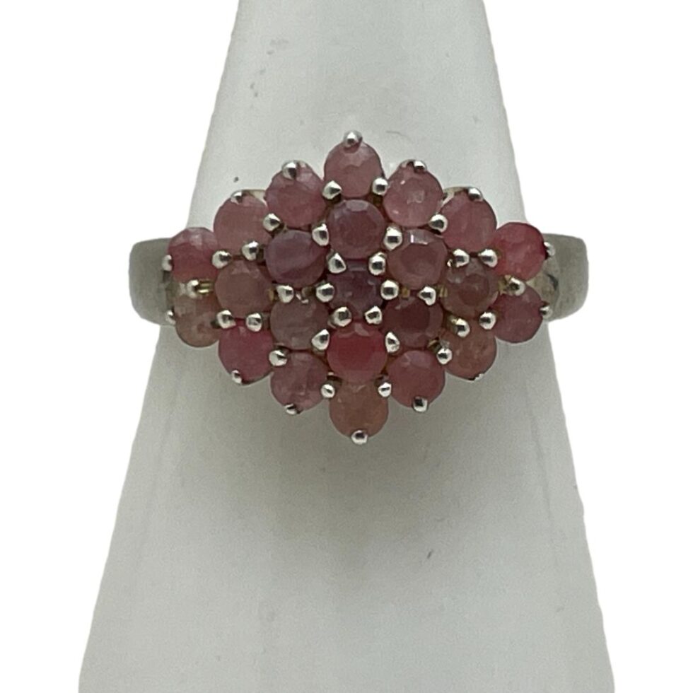 Main photo of silver ruby cluster ring displayed on a white cone shaped stand and seen with the ring front forward facing. This is quite an unusual ring in that the cluster is side wide rather than circular. The rubies are a sweet pale pink and red colour mix which marries well with the silver.