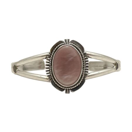 Main photo of Navajo signed cuff bracelet displayed on a flat surface against a white background with the front of bracelet at centre and forward facing. The cuff has a split band at front with an oval central frame which houses the dusky pink coloured natural shell. The frame is a very recognisable Navajo design with 'feathered' edges at the 4 compass points and intricate rope twist around the stone.
