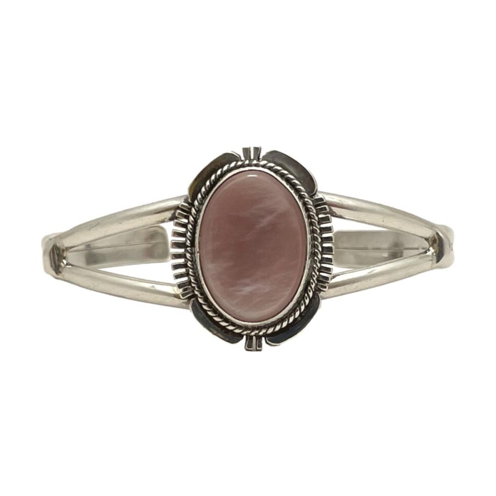 Main photo of Navajo signed cuff bracelet displayed on a flat surface against a white background with the front of bracelet at centre and forward facing. The cuff has a split band at front with an oval central frame which houses the dusky pink coloured natural shell. The frame is a very recognisable Navajo design with 'feathered' edges at the 4 compass points and intricate rope twist around the stone.
