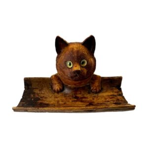 Main photo of Black Forest cat inkwell seen from an eye level from the front so the cat is looking straight at you! The cats head holds the inkwell and from nose up is the lid cover that disguises the ink pot. The cat has yellow glass eyes that seem to stare right at you. in front of the cats face is a curved piece of wood - a little like bamboo split in half where the pen or quill would rest. The cats has its small front paws over the top of the side of the pen rest appearing as though it is peering over the edge.