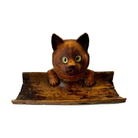 Main photo of Black Forest cat inkwell seen from an eye level from the front so the cat is looking straight at you! The cats head holds the inkwell and from nose up is the lid cover that disguises the ink pot. The cat has yellow glass eyes that seem to stare right at you. in front of the cats face is a curved piece of wood - a little like bamboo split in half where the pen or quill would rest. The cats has its small front paws over the top of the side of the pen rest appearing as though it is peering over the edge.
