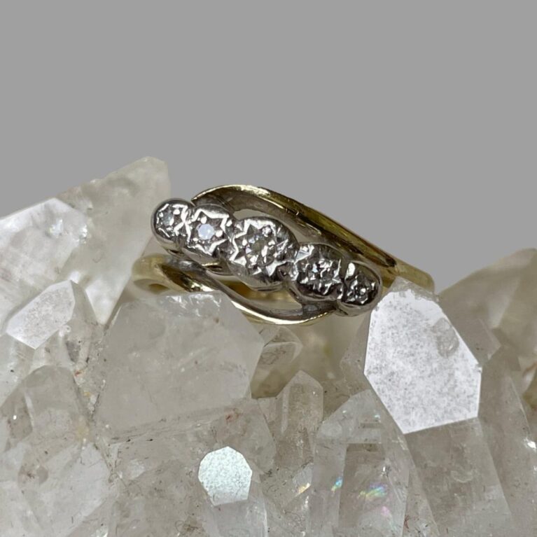 Main photo of gold platinum diamond ring displayed on top of a rock of natural quartz crystal and seen with ring front facing forward. The gold band is split with each end curved over the diamonds which are set in platinum at the centre. The ring is displayed on top of a rock of natural quartz crystal.