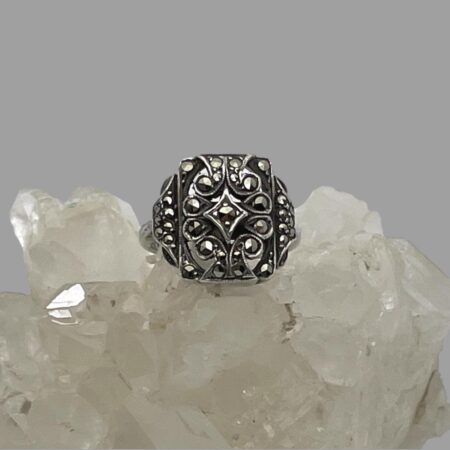 Main photo of adjustable silver marcasite ring displayed on top of a rock of natural quartz crystal with the ring front forward facing. The ring has a minimalist design of a squarish front with a small number of marcasite pieces set within. At the front, each marcasite has its own silver setting including a diamond shaped central frame also with marcasite.