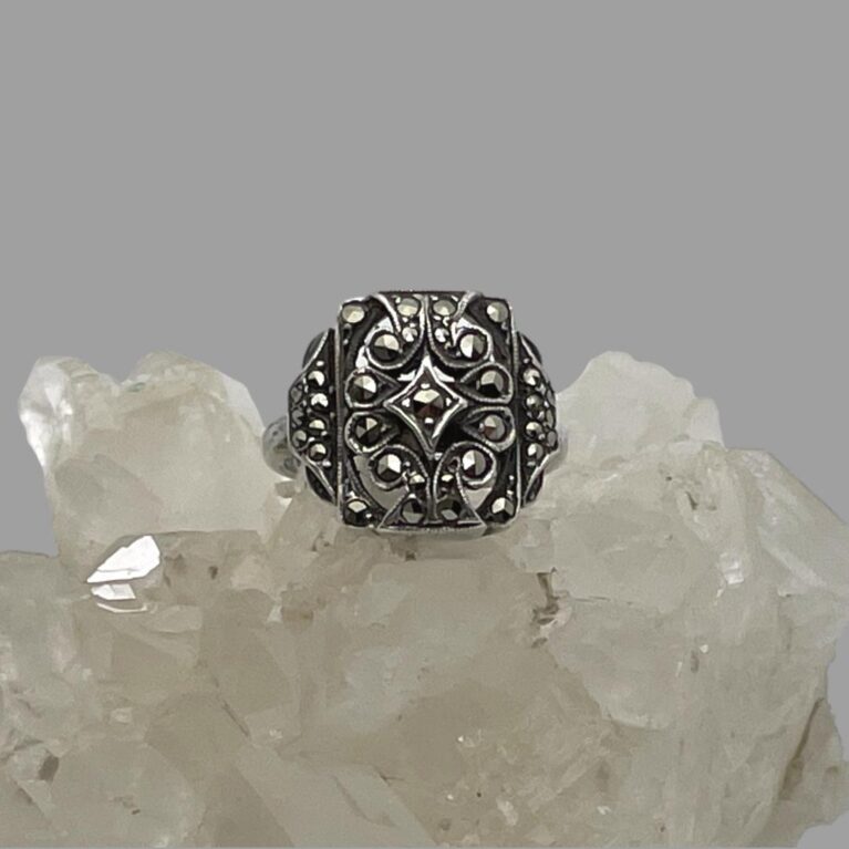 Main photo of adjustable silver marcasite ring displayed on top of a rock of natural quartz crystal with the ring front forward facing. The ring has a minimalist design of a squarish front with a small number of marcasite pieces set within. At the front, each marcasite has its own silver setting including a diamond shaped central frame also with marcasite.