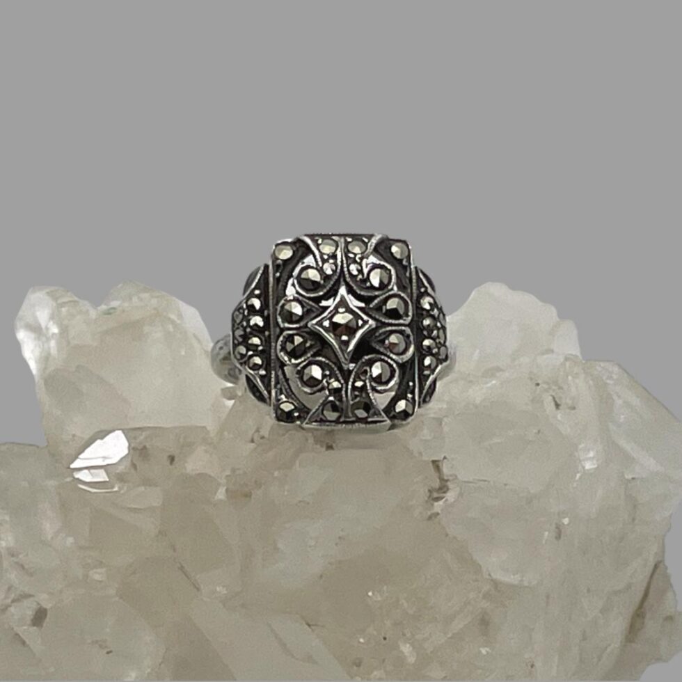 Main photo of adjustable silver marcasite ring displayed on top of a rock of natural quartz crystal with the ring front forward facing. The ring has a minimalist design of a squarish front with a small number of marcasite pieces set within. At the front, each marcasite has its own silver setting including a diamond shaped central frame also with marcasite.
