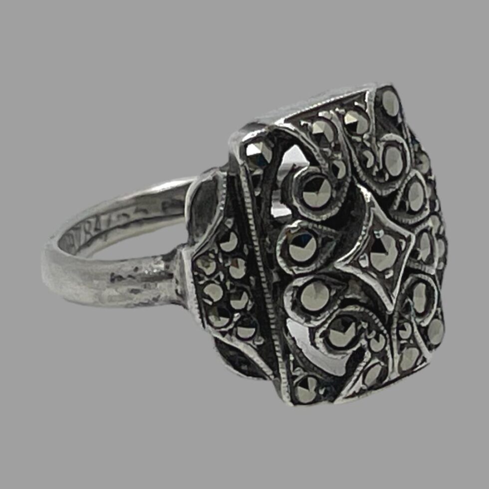 Photo of adjustable silver marcasite ring displayed on a flat surface with ring front facing towards the right corner. The extra section by the ring shoulders which has some marcasite is visible.