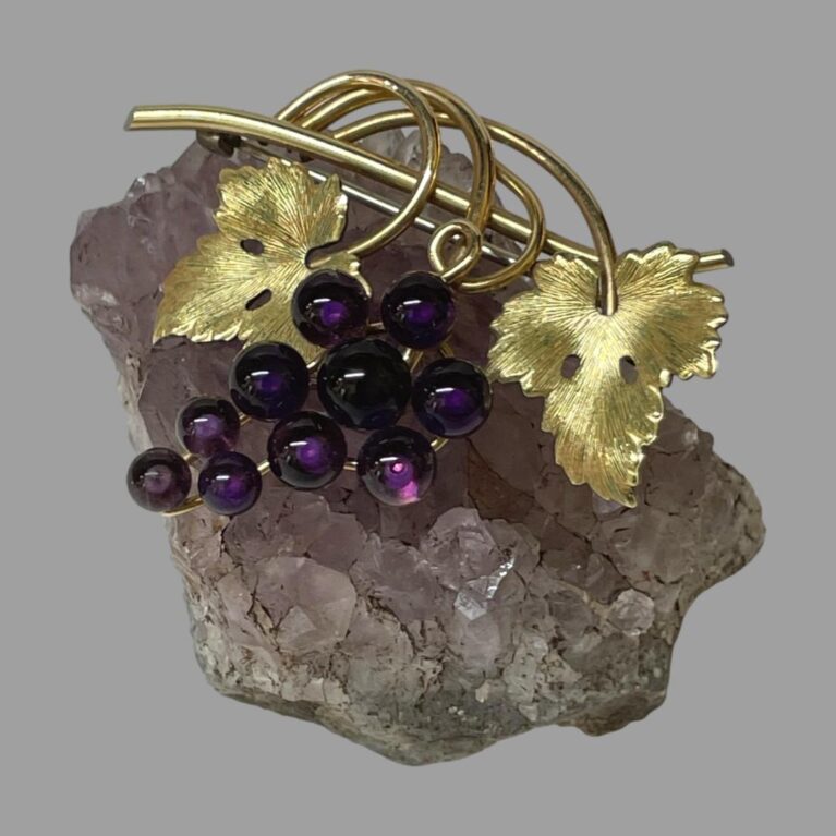 Main photo of signed amethyst grapes brooch displayed balanced on top of a small rock of amethyst. The grape bunch is made up of 10 round amethyst stones with larger ones at top graduating down to smallest at the tip. The amethyst colour is a deep purple. The bar of the brooch is the stalk which has vine coiled around it with a textured vine leaf on either side of the grapes. The gilt of the stalk, vine and leaves and the deep purple of the grapes compliment each other perfectly. A wonderful gift for a wine lover!