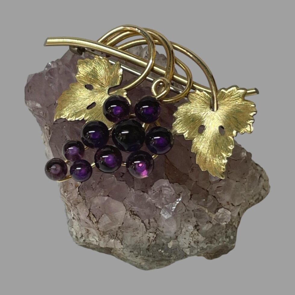 Main photo of signed amethyst grapes brooch displayed balanced on top of a small rock of amethyst. The grape bunch is made up of 10 round amethyst stones with larger ones at top graduating down to smallest at the tip. The amethyst colour is a deep purple. The bar of the brooch is the stalk which has vine coiled around it with a textured vine leaf on either side of the grapes. The gilt of the stalk, vine and leaves and the deep purple of the grapes compliment each other perfectly. A wonderful gift for a wine lover!