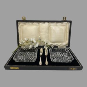Main photo of cased butter dishes &knives displayed inside their original fitted case. The lead crystal butter dishes are to the left and right inside the case with the small silver butter knives slotted vertically at the centre. The case is in very good condition except for mild foxing of the silk to inside lid. A spray of white orchids is between the lid silk and fitted velvet area for display purposes only.