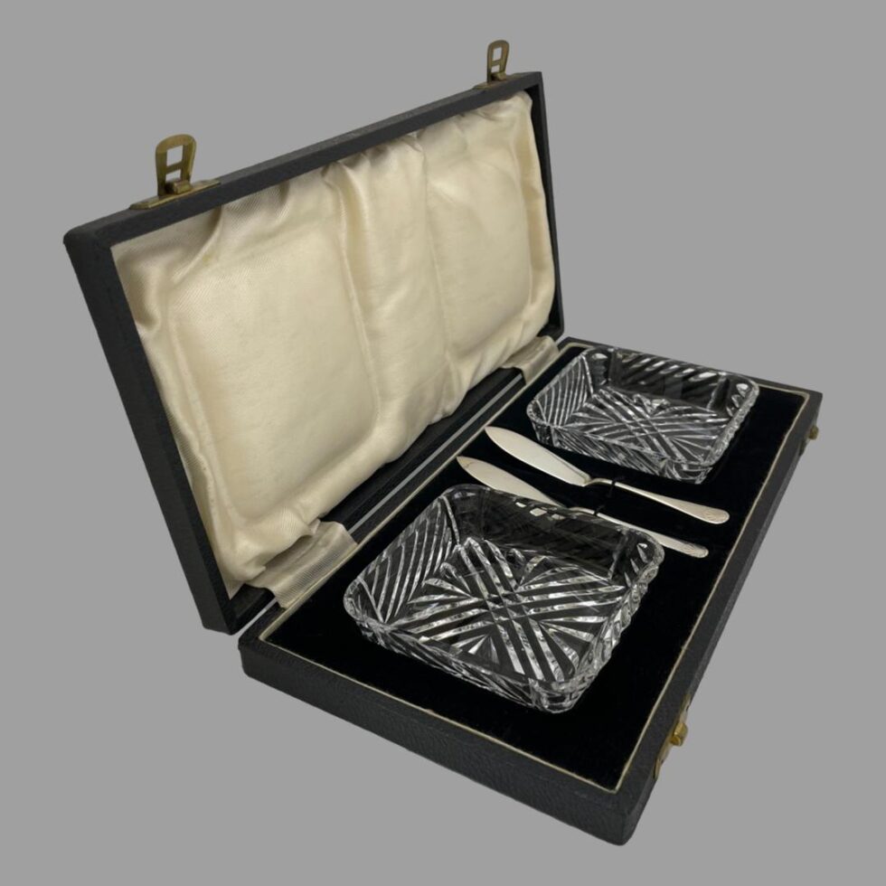 Photo of cased butter dishes &knives displayed inside their fitted case facing sideways to the right.