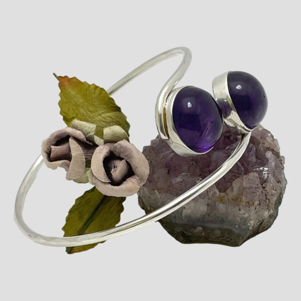 Photo of Modernist silver amethyst bracelet still displayed with the cabochons end resting on the rock of natural amethyst this time shown from a side angle. The posy of faux lilac roses is still to the left.