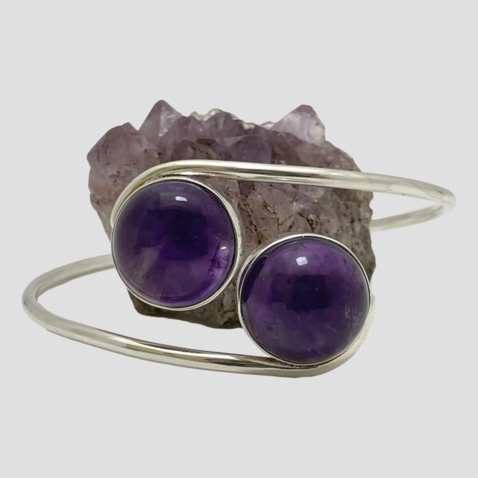Photo of Modernist silver amethyst bracelet displayed on a flat surface with the amethyst fronts in the foreground. The small rock of natural amethyst has been placed in the centre of the band immediately behind the amethyst cabochons which are a darker richer shade of purple.