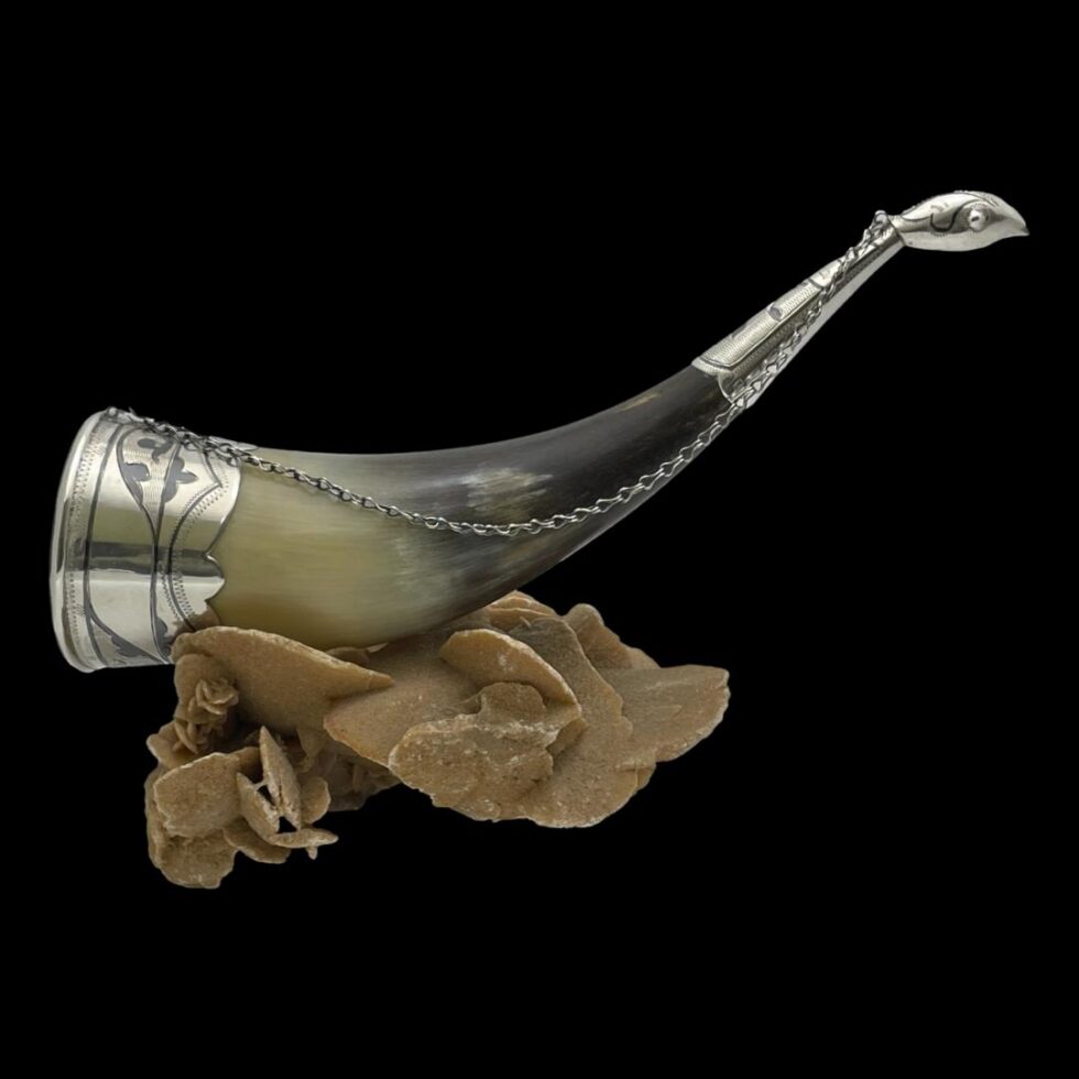 Main photo of Russian horn drinking vessel displayed on top of a rock of natural desert sand rose. The pointed tip of the horn has a little silver covering which has a birds head at the end. The mouth end of the horn has a one and a half inch silver rim on the outside. A silver chain extends from the mouth of the vessel to the back of birds head at the tip making a handy carrying handle that can be hooked to a belt or hung up to dry after use. A perfect gift for the Viking in your life!