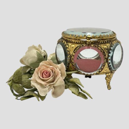 Main photo of antique French bijouterie casket displayed from a near eye level with one of the side 'windows' in the foreground and showing the padded red silk interior. There is a small posy of pink faux roses to the left for display only.