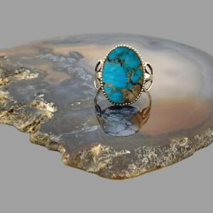 Main photo of antique gold turquoise ring displayed on top of a slice of natural agate stone with the ring front forward facing. The turquoise has a lovely colouring with natural stone inclusions giving it a very rustic natural look. The gold frame around the ring appears like tiny teeth holding the stone in place and pretty unusual 'wing' like shoulders.