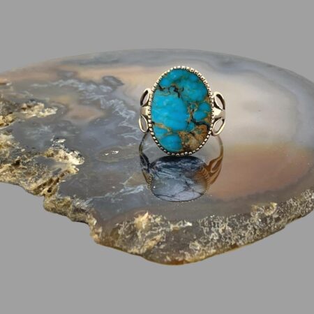 Main photo of antique gold turquoise ring displayed on top of a slice of natural agate stone with the ring front forward facing. The turquoise has a lovely colouring with natural stone inclusions giving it a very rustic natural look. The gold frame around the ring appears like tiny teeth holding the stone in place and pretty unusual 'wing' like shoulders.