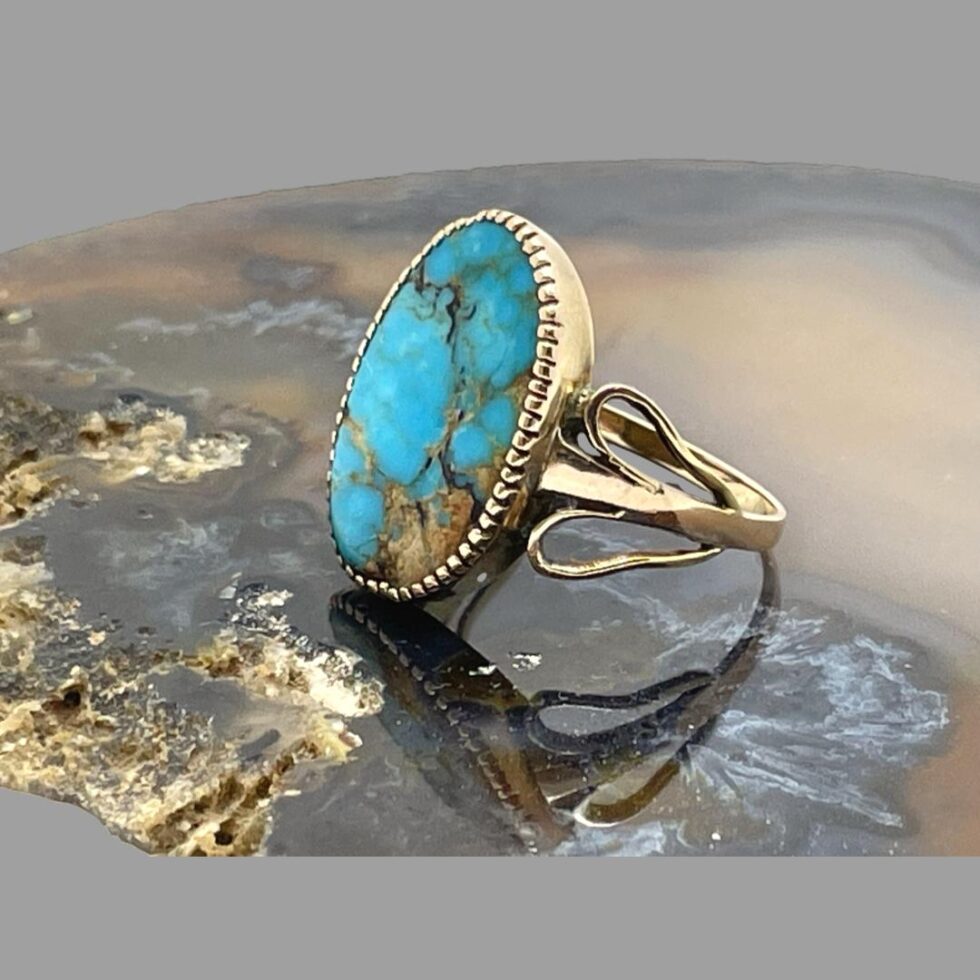 Photo of antique gold turqupoise ring still displayed on top of a slice of agate this time with the ring front faced to the left. The unusual wing like shoulders are visible - it is formed by 2 short looped bands which are added to the main ring band. The tips of the wings, the wider end, make the shoulders.