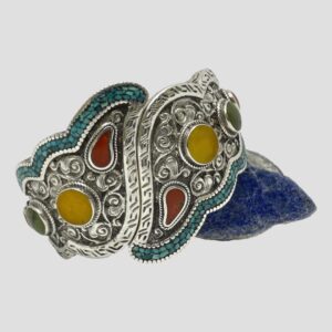 Main photo of chunky hinged silver bracelet displayed leaning on a small rock of lapis on one side. The front of the bracelet is on display. There is a lot of detailing on the silver areas with different styles of engravings throughout. The mosaic turquoise is laid along one edge of opposing sides and the colourful stones are set in different shapes within their own frames. The stones are bright red, yellow and forest green adding a joyful colour uplift with the turquoise. A true statement piece - be prepared for many compliments!